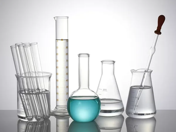 Glass test tubes and flasks with laboratory liquids are arranged beside a dropper on a reflective surface.