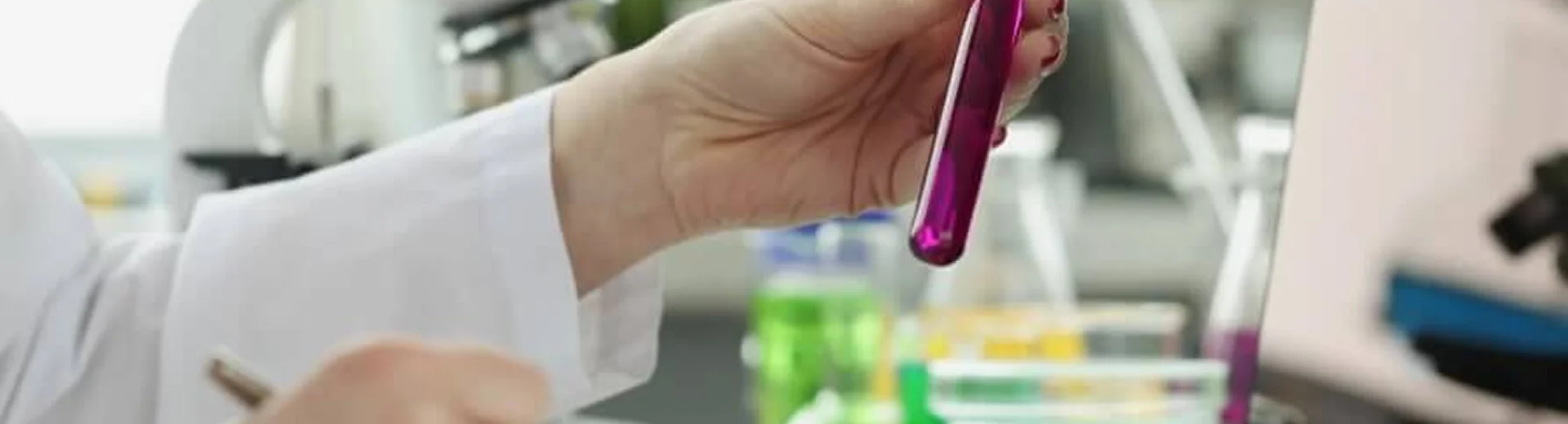 A researcher records results for a test tube containing purple reagent beside a microscope and laptop.