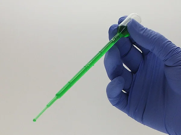 A gloved lab technician uses a Pasteur pipette to transfer green liquid.