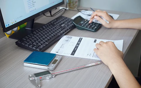 An employee uses a calculator to prepare a quote, with documents and a digital caliper on the desk.